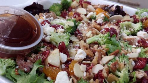 A colorful salad with mixed greens, broccoli, almonds, cranberries, feta cheese, and a side of dressing.