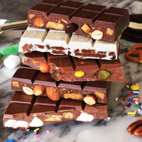 A stack of colorful chocolate bars with various toppings, including nuts, candies, and marshmallows, on a marble surface.
