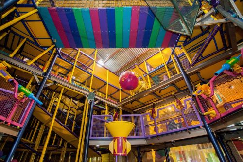 Colorful indoor play structure with multiple levels, slides, and climbing areas, featuring a large red ball at the center.