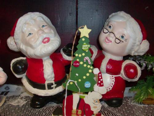 Two cheerful Santa and Mrs. Claus figurines stand beside a small decorated Christmas tree.