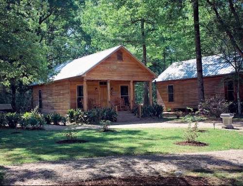 Two wooden cabins surrounded by lush greenery and trees, with a gravel path leading to the entrance.