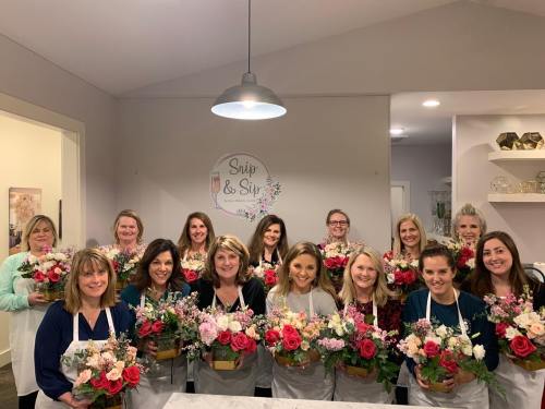 A group of women holding flower arrangements, smiling, in a bright room with a "Snip & Sip" sign on the wall.