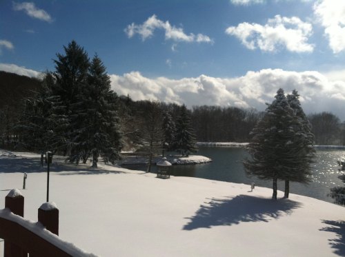 A snowy landscape with trees and a calm lake under a bright blue sky with fluffy clouds.