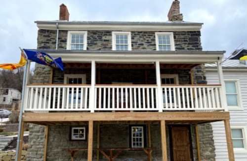 A two-story stone house with a wooden porch, featuring flags and a cloudy sky in the background.