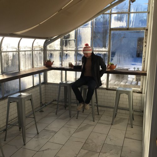 A person sits on a stool in a bright, glass-enclosed space with plants, wearing a striped hat and holding a drink.