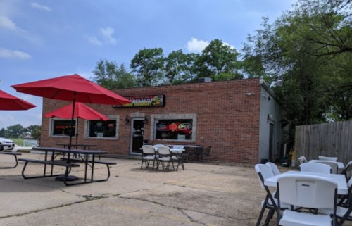 A brick restaurant with outdoor seating under red umbrellas, surrounded by trees and a clear blue sky.