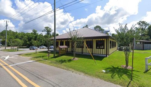 A yellow and brown building with a metal roof, surrounded by greenery, located at a street intersection on a sunny day.
