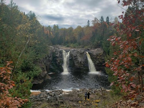 A scenic view of a waterfall surrounded by autumn foliage and a person standing near the water's edge.