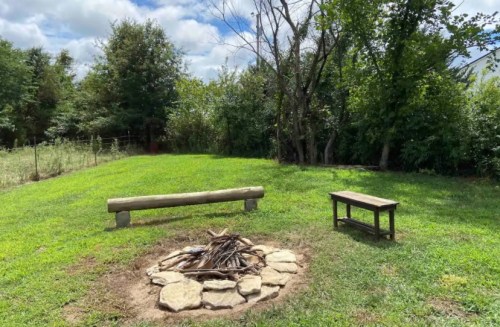 A grassy area with a fire pit made of stones, surrounded by trees, and a wooden bench nearby under a cloudy sky.