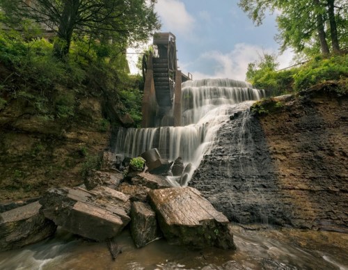 A serene waterfall cascades over rocks, with a historic mill structure in the background surrounded by lush greenery.