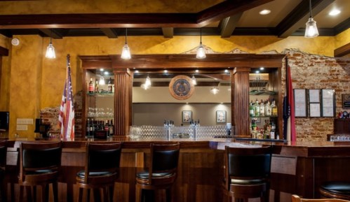 A cozy bar with wooden stools, a well-stocked counter, and warm yellow walls, featuring an American flag and decorative lighting.
