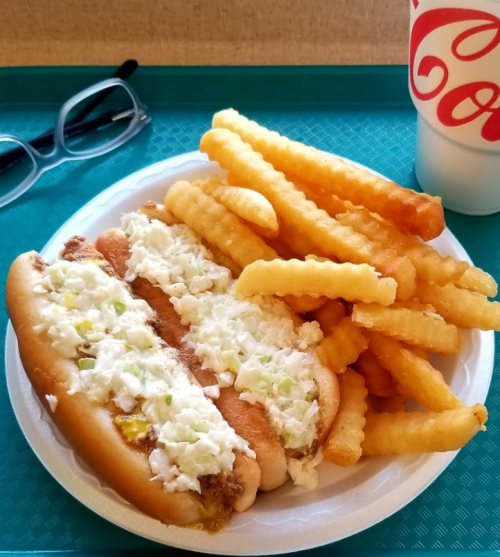A plate with two hot dogs topped with coleslaw, served with crinkle-cut fries and a cup of soda on the side.