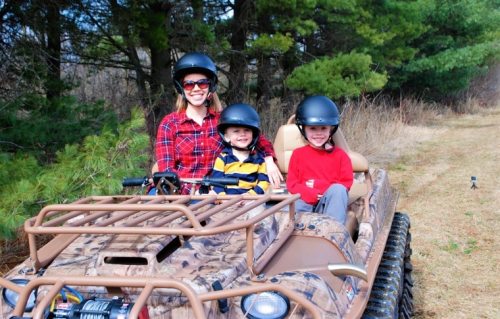 A woman and two children wearing helmets sit in an all-terrain vehicle on a grassy path surrounded by trees.