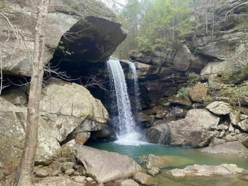 A serene waterfall cascades over rocks into a clear pool, surrounded by lush greenery and rugged stone formations.