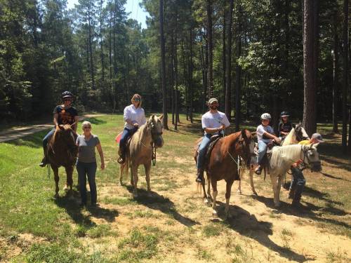 A group of six people on horseback in a wooded area, with two individuals standing beside the horses.