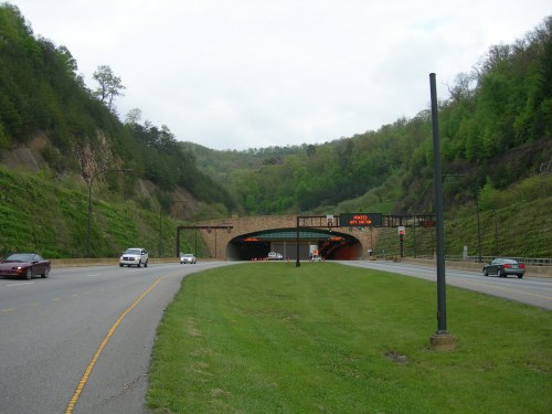 A highway leading to a tunnel surrounded by green hills and trees, with cars approaching the entrance.