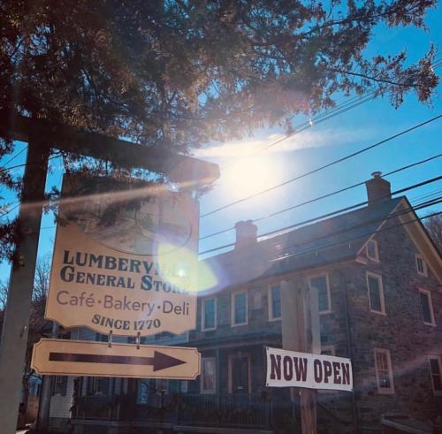 Sign for Lumberville General Store, indicating it's open, with a sunny sky and historic buildings in the background.