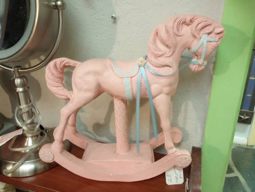 A pink rocking horse figurine with blue accents, displayed on a wooden shelf next to a mirror.