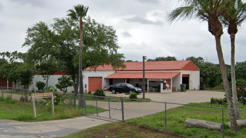 A large white building with a red roof, surrounded by palm trees and a fenced area, with a car parked outside.