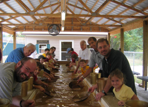 A group of children and adults panning for gold at a covered outdoor facility, smiling and engaged in the activity.