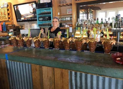 A bartender stands behind a bar with a row of large shrimp cocktails garnished with fries and colorful straws.
