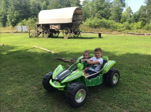 Two children ride a green toy vehicle on grass, with a wagon in the background and trees surrounding the area.