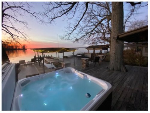 A hot tub on a deck overlooking a lake at sunset, with trees and outdoor seating in the background.