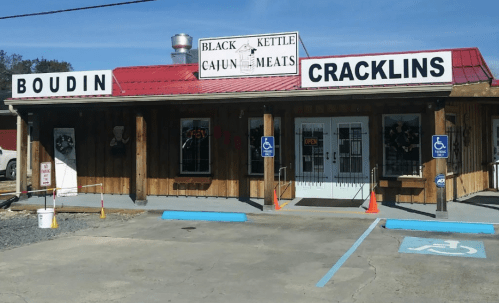 A rustic storefront with signs for "Boudin" and "Cracklins," featuring a red roof and accessible parking.