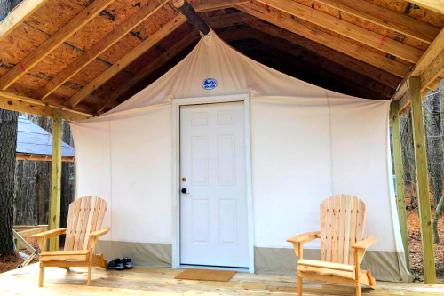 A rustic cabin with a white canvas exterior, a door, and two wooden chairs on a porch surrounded by trees.