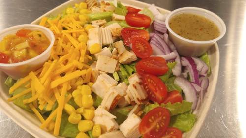 A colorful salad with lettuce, tomatoes, corn, shredded cheese, chicken, red onion, and two dressings on the side.