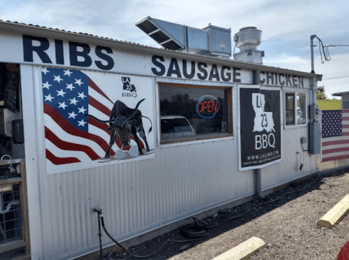 A BBQ food stand with an American flag mural, featuring signs for ribs, sausage, and chicken.