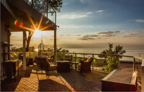 A serene patio with chairs and a table, overlooking a lake at sunset, with warm sunlight filtering through trees.