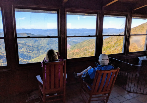 Two people sit in rocking chairs by a large window, enjoying a scenic mountain view.