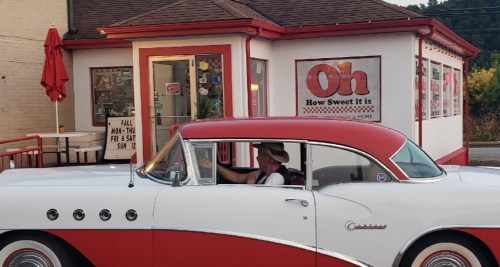A classic red and white car drives past a retro-style ice cream shop with a red umbrella outside.
