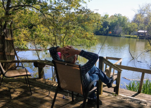 A person relaxes in a chair by a serene lake, with hands on their head, surrounded by trees and nature.