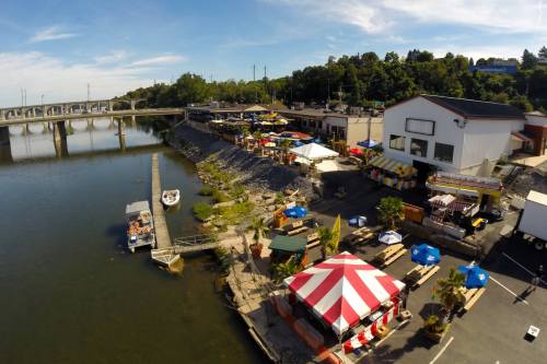 A riverside scene featuring colorful tents, boats, and a restaurant along the water, with a bridge in the background.