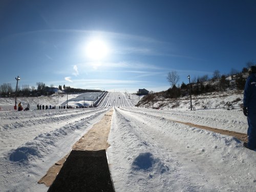 A snowy hill with multiple sledding lanes under a bright sun, surrounded by trees and a clear blue sky.