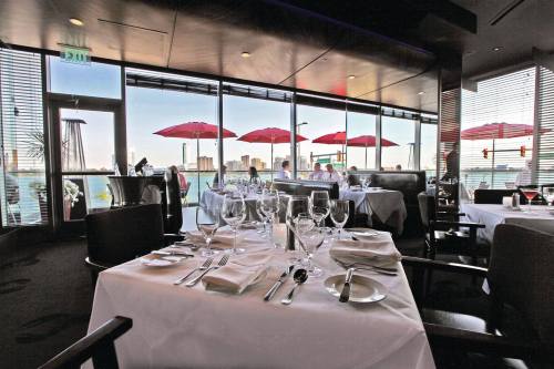 Elegant restaurant setting with tables set for dining, large windows showcasing a city view and red umbrellas outside.