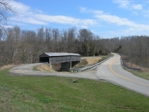 A rustic covered bridge spans a small stream, surrounded by trees and a winding road under a blue sky.
