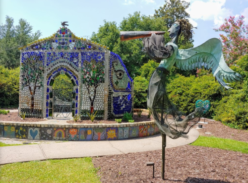 A colorful mosaic structure with a glass bottle design and a metal angel sculpture holding a telescope in a garden.