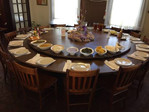 A large round dining table set with empty plates and bowls of various dishes, surrounded by wooden chairs.