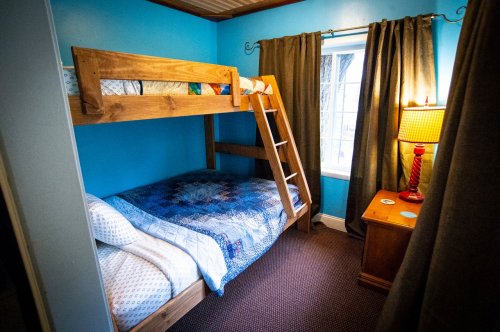 A cozy bedroom with a blue wall, a bunk bed, a single bed, curtains, and a lamp beside a window.