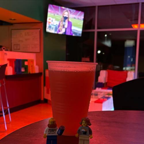 A drink in a cup sits on a table with LEGO figures, while a sports event is shown on a TV in the background.