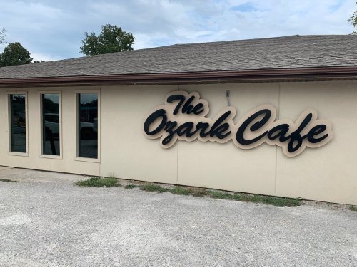 Signage of "The Ozark Cafe" on a beige building with large windows and a gravel parking area.
