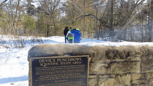 A group of people in winter clothing near a stone sign for Devil's Punchbowl Scientific Study Area, surrounded by snow.