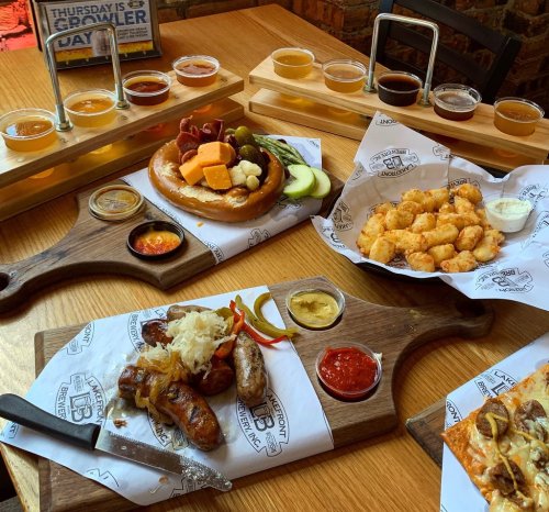 A wooden table with various food platters, including sausages, cheese, pretzels, tater tots, and beer flight samples.