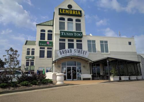 A multi-story building featuring signs for Lemuria, Tuxes Too, and Fresh Ink, with outdoor seating and a clear sky.