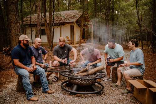 A group of six men gathered around a fire pit in a wooded area, enjoying drinks and conversation. A cabin is in the background.