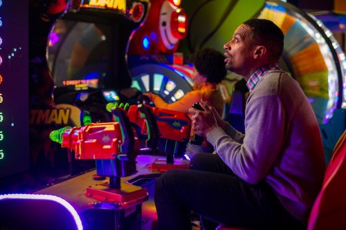 A man plays an arcade shooting game, focused and holding a bright red controller, with colorful lights in the background.