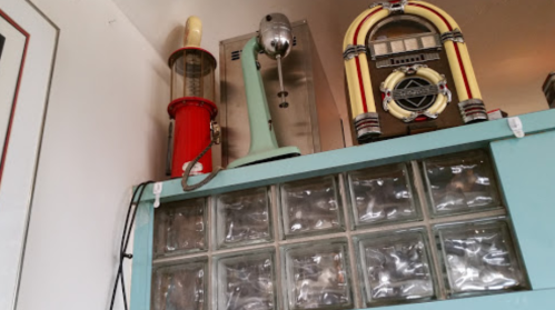 A vintage kitchen display featuring a red soda fountain, green mixer, and a retro jukebox on a glass block wall.